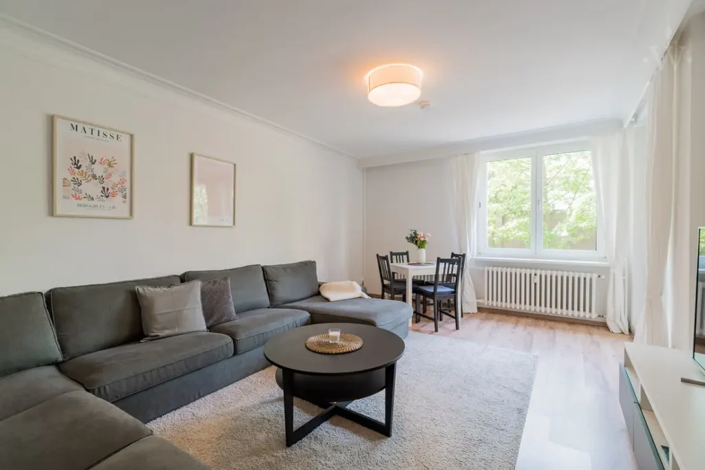 Bright living room with grey corner sofa, round coffee table and dining area by the window.