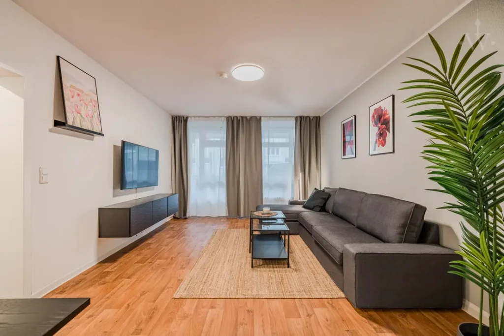 Modern living room with grey corner sofa, wooden floor, TV on the wall, decorative pictures and large windows with curtains.