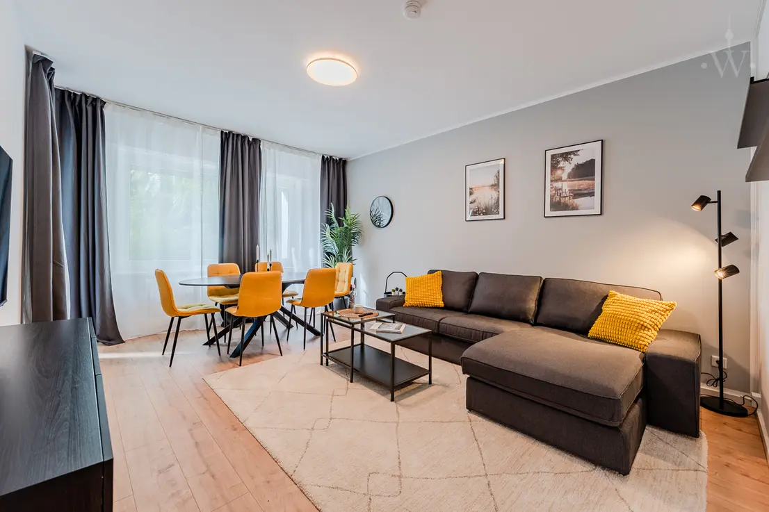 Wohnzimmer mit grauer Eckcouch, gelbem Essbereich und großen Fenstern, moderne Wandbilder.