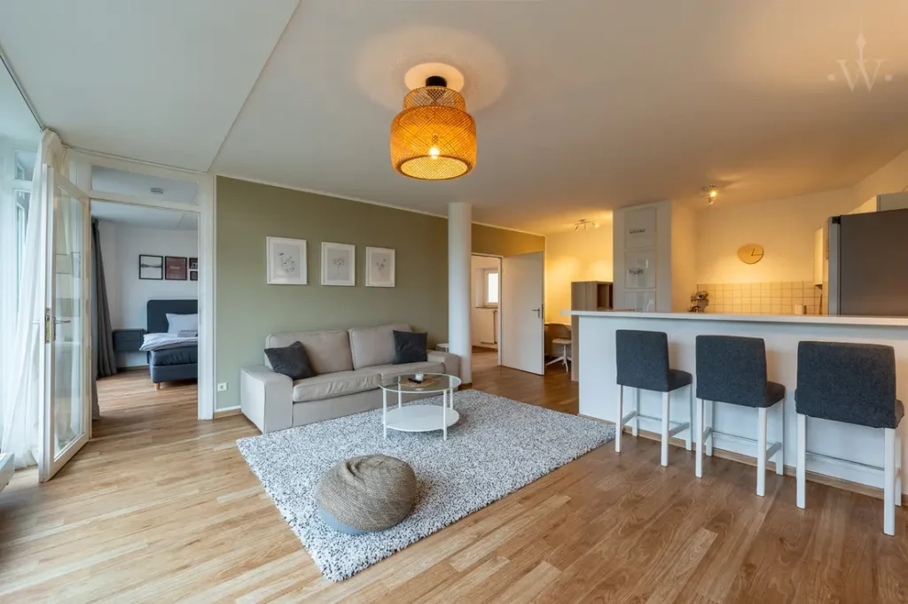 Eine Couch mit einem kleines Tisch und einem Teppich. Ebenfalls sind da 3 Stühle zu sehen und ein Teil der Küche