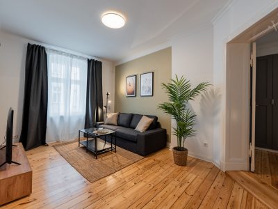 Helles Wohnzimmer mit grauem Sofa, Holzboden und großer Zimmerpflanze nahe dem Fenster